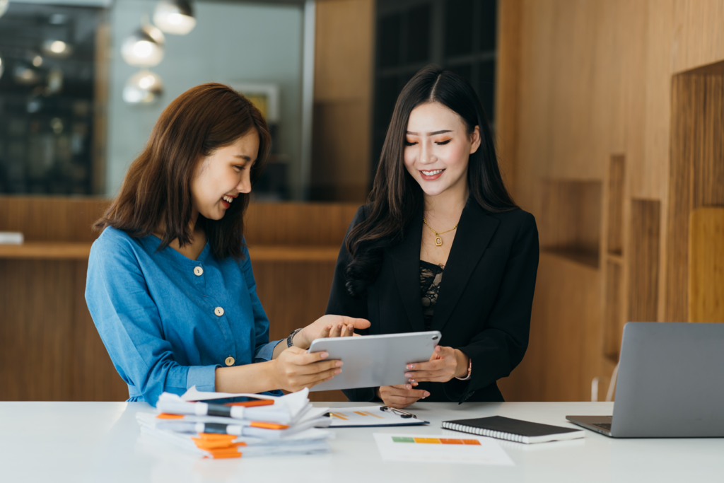 Two Asian businesswomen collaborating on a tablet in a modern office setting.